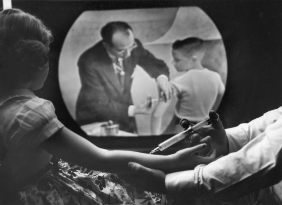 As eight-year-old girl receives a polio vaccine, she watches a closed circuit television broadcast (showing Dr Jonas Salk as he inoculates a boy) of a training telecast for physicians and scientists, New York, New York, April 12, 1955.