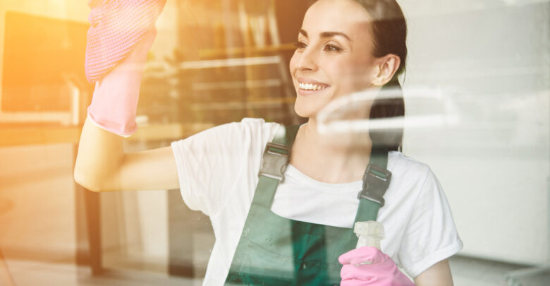 beautiful smiling young woman cleaning and wiping window with spray bottle and rag